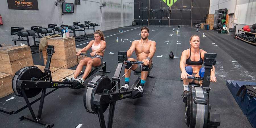 Três atletas fazendo exercício de remo indoor em uma box de CrossFit, focados na performance e resistência.