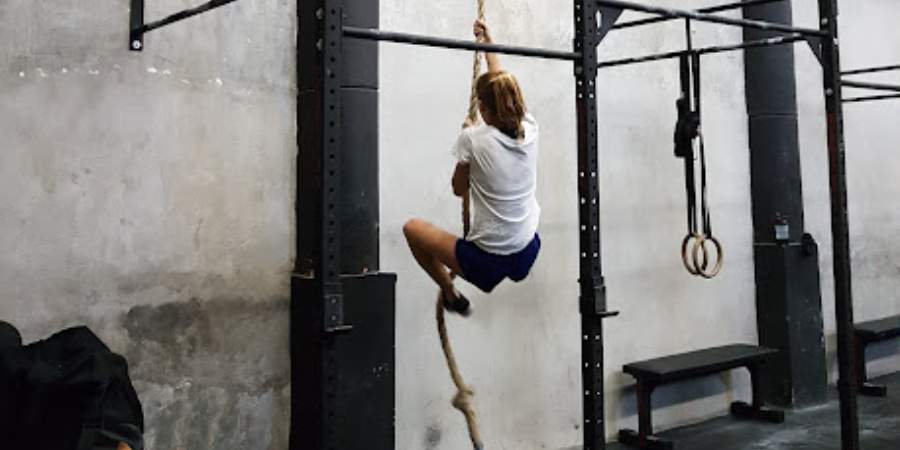 Atleta praticando escalada de corda em box de CrossFit, destacando habilidade e treino de força funcional.