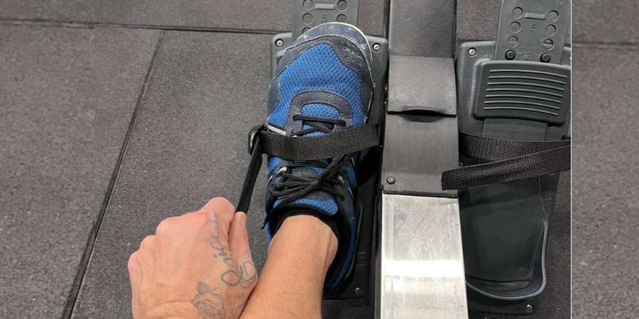 Um atleta de CrossFit com tatuagem no braço ajusta o strap sobre seu tênis azul em um ergômetro de remo, preparando-se para uma sessão de treino intensa.
