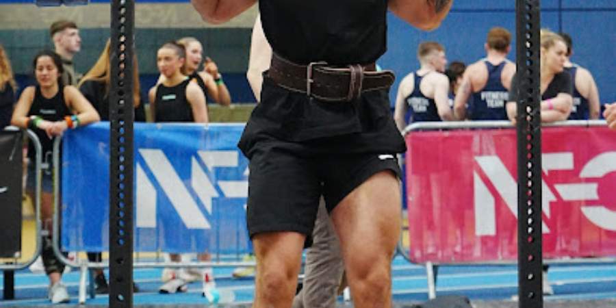 Um atleta de frente para a câmera prepara-se para realizar um deadlift em um evento de CrossFit, com um cinto de levantamento para suporte, enquanto espectadores ao fundo observam atentamente.