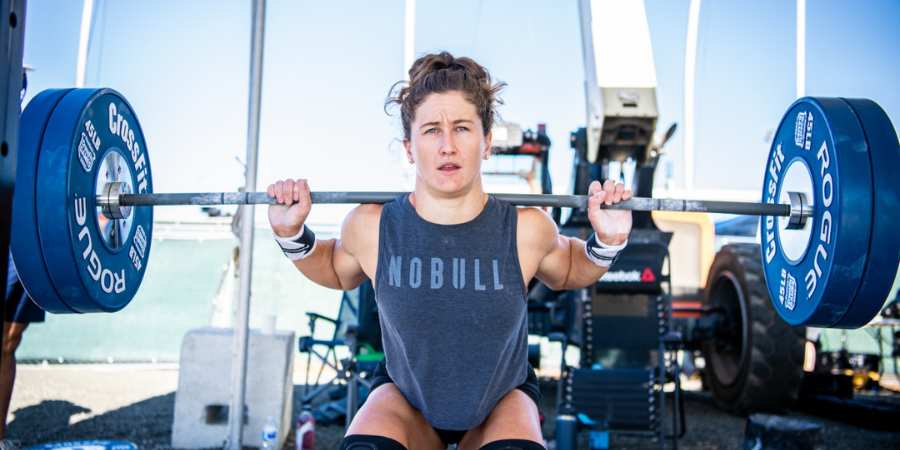 Uma atleta feminina de CrossFit concentrada prepara-se para realizar um back squat, segurando uma barra carregada com pesos Rogue, com um cenário ao ar livre e equipamentos de treino ao fundo, ilustrando determinação e força no levantamento de peso.