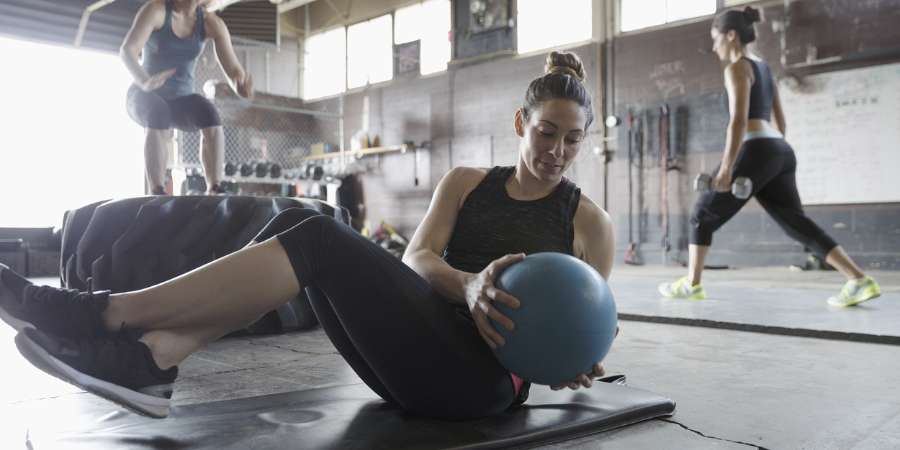 Uma mulher pratica exercícios de core com uma medicine ball em uma academia de CrossFit, enquanto outras pessoas realizam exercícios de agachamento e corrida ao fundo, destacando o ambiente dinâmico e variado do treinamento funcional.
