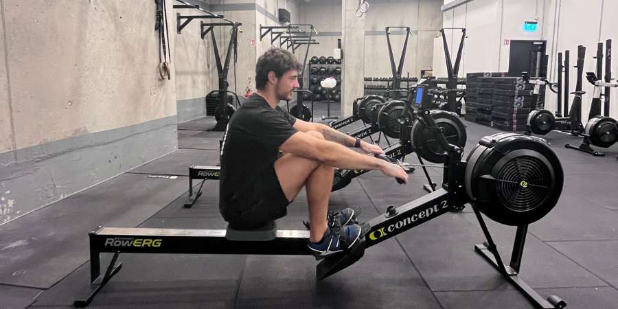 Homem treinando em um ergômetro Concept2 RowErg em uma academia de CrossFit, focando na técnica e na performance de remo indoor.