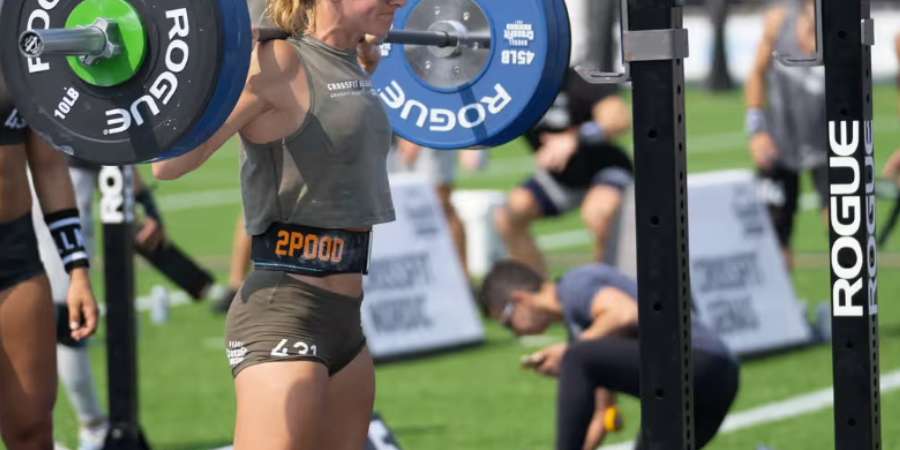 Uma atleta de CrossFit focada realiza um back squat com pesos Rogue, usando um cinto de levantamento de peso para suporte, em um ambiente de competição ao ar livre, com outros competidores ao fundo.