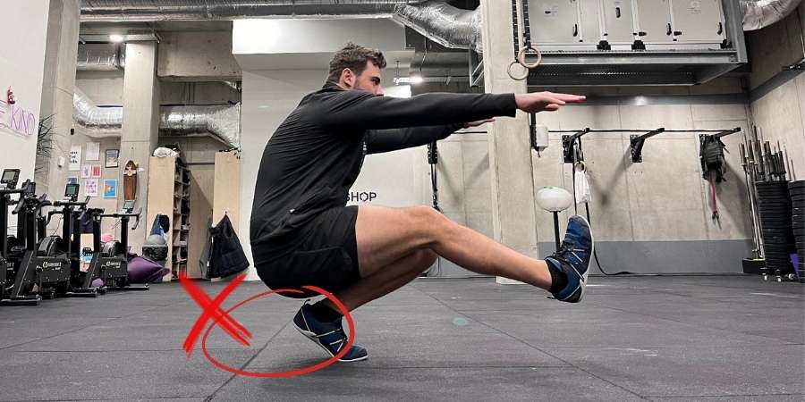 Um homem pratica o exercício pistol squat em uma academia de CrossFit, com um "X" vermelho indicando uma técnica incorreta na posição do pé, em um ambiente equipado para treino funcional.