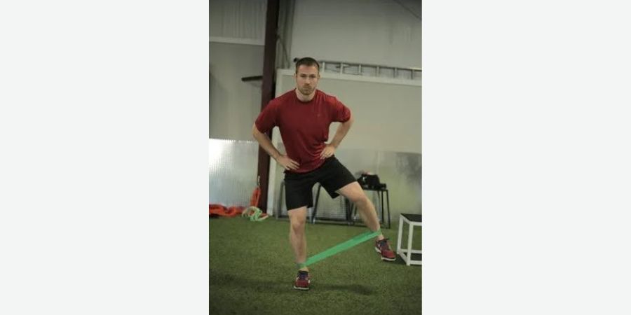 Um homem em uma camiseta vermelha e shorts realiza um exercício de estabilidade do quadril com uma banda de resistência verde, em uma academia com grama artificial, focando na melhoria da força lateral e mobilidade.