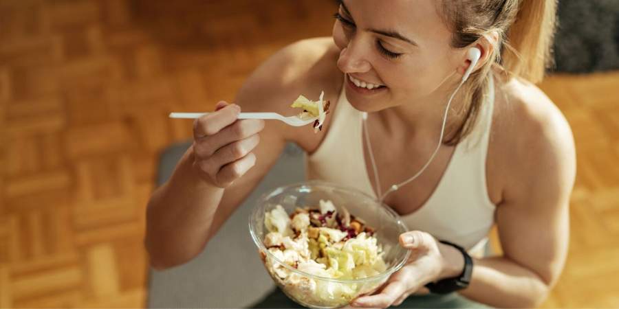 Mulher sorridente com fones de ouvido comendo uma salada saudável, representando a importância da nutrição balanceada em conjunto com um estilo de vida ativo e fitness.