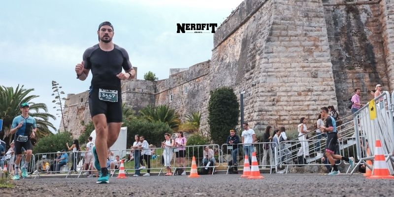 Hugo Casarin corre durante uma prova de rua, vestindo roupa esportiva preta e tênis azul. Ele aparece concentrado no percurso, com sua numeração presa na cintura. Ao fundo, há um forte de pedra histórico, espectadores observando atrás das grades e outros atletas no trajeto. O logo “NerdFit” está centralizado na parte superior da imagem.