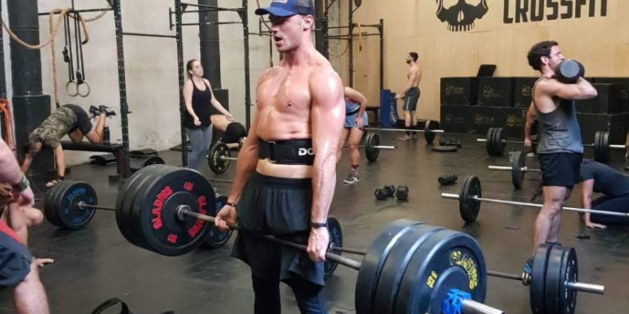 Atleta masculino realiza levantamento de peso em uma academia de CrossFit repleta de praticantes, demonstrando força e concentração em meio a uma sessão intensa de treinamento, com o logotipo do CrossFit visível ao fundo.