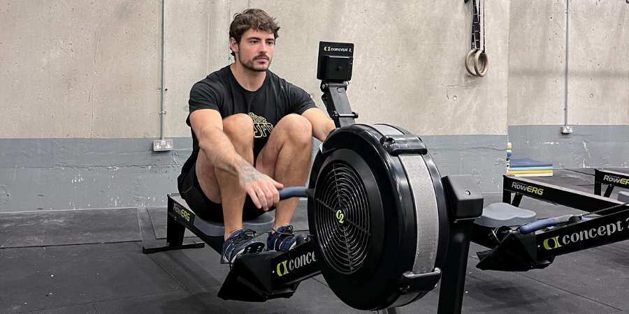 Um atleta de CrossFit concentrado realiza um treino de remo indoor em um ergômetro Concept2, aprimorando sua resistência cardiovascular e força muscular em um ambiente de box de CrossFit.