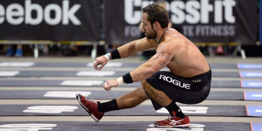 Um atleta masculino de CrossFit realiza um pistol squat durante uma competição, destacando-se com seu equipamento de levantamento de peso e o logotipo da Reebok ao fundo, simbolizando foco e força em condições de alta pressão.