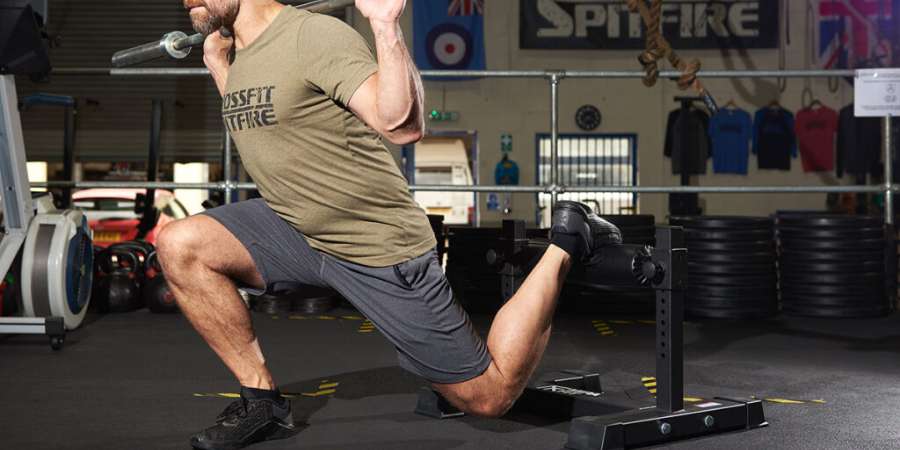 Um homem realiza uma variação de agachamento com um joelho apoiado em uma academia de CrossFit, vestindo uma camiseta com a logo da CrossFit e calças esportivas, ilustrando a força e a técnica em um ambiente de treino funcional.